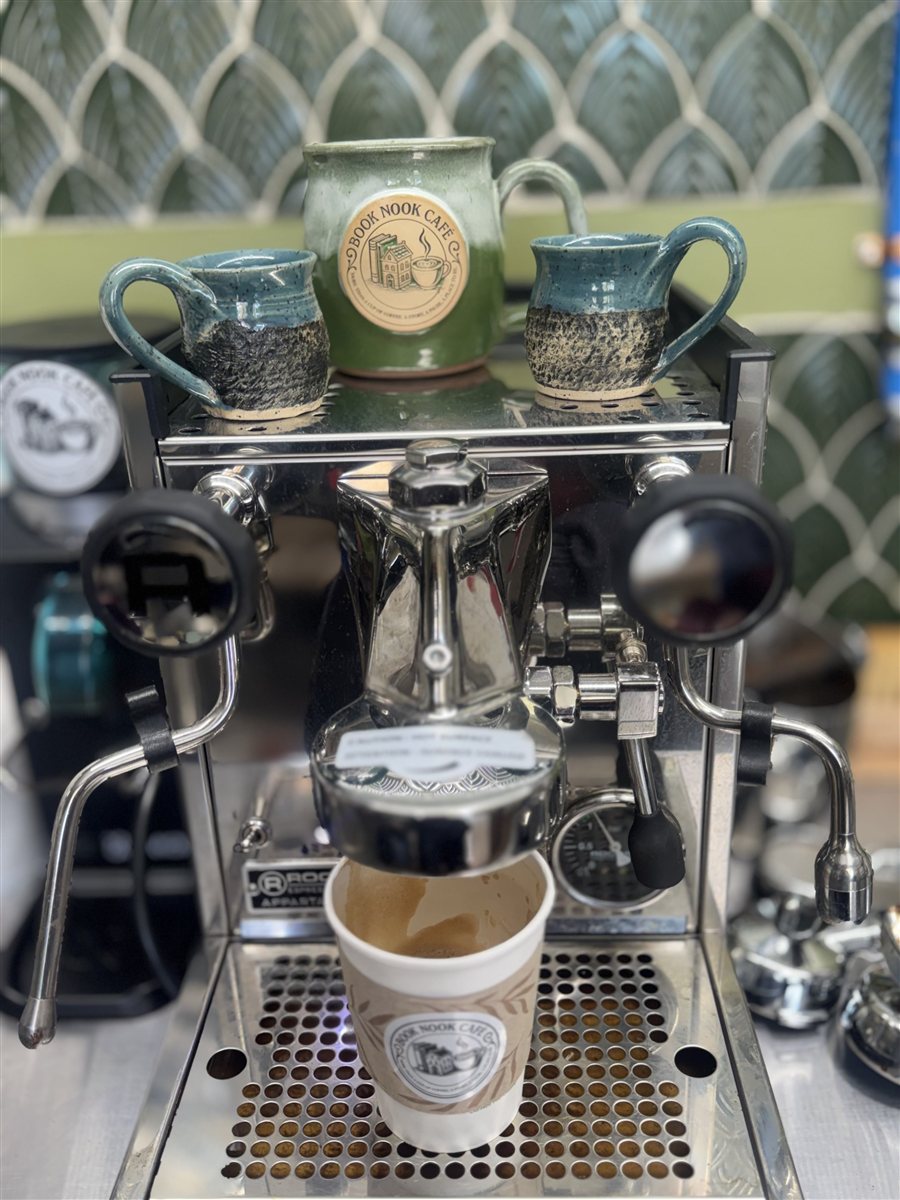 Espresso machine pouring coffee into a Book Nook Cafe cup, with ceramic mugs on top.