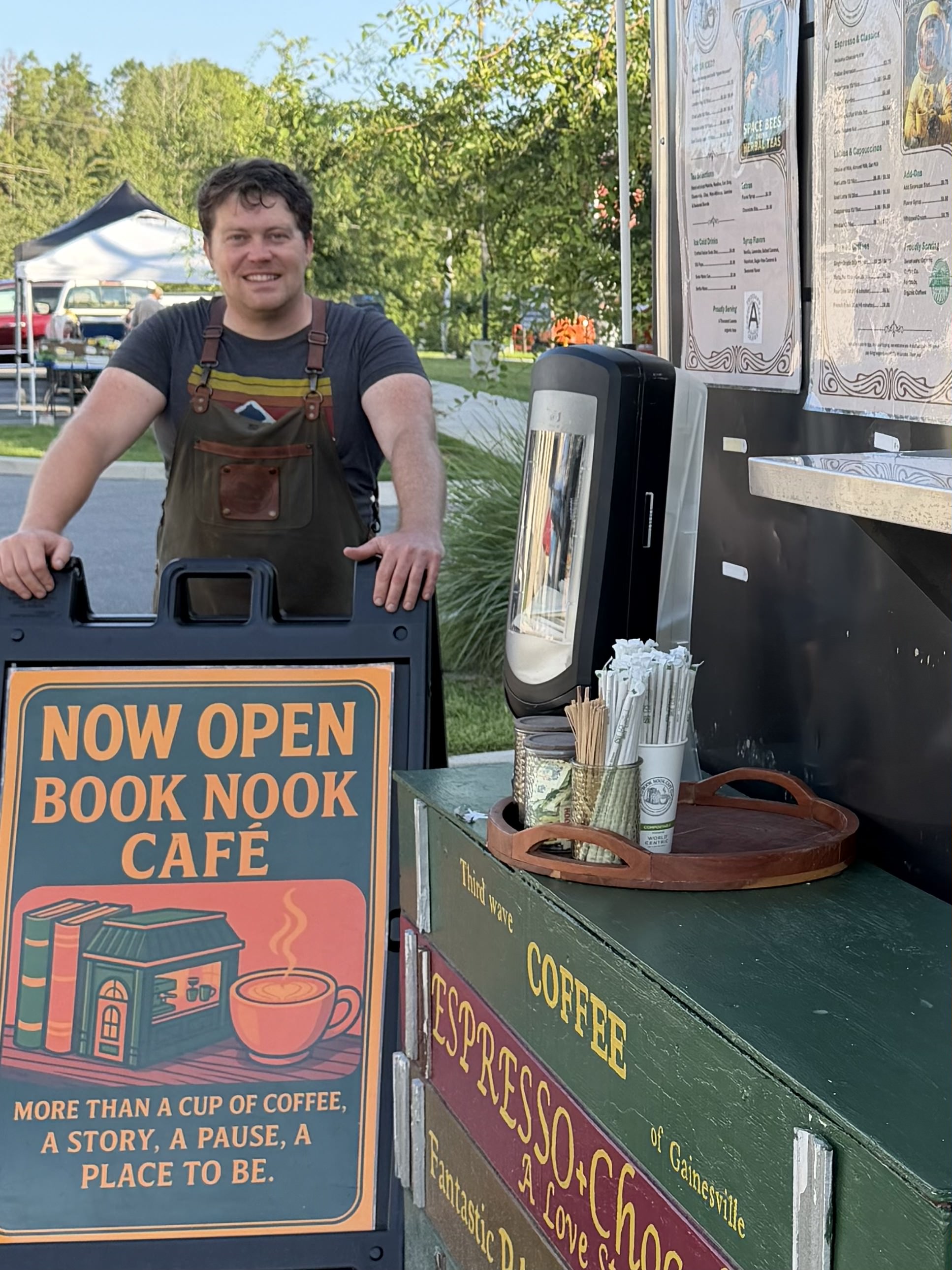 A smiling barista in an apron beside the Book Nook Cafe trailer and its Now Open sign in Gainesville.