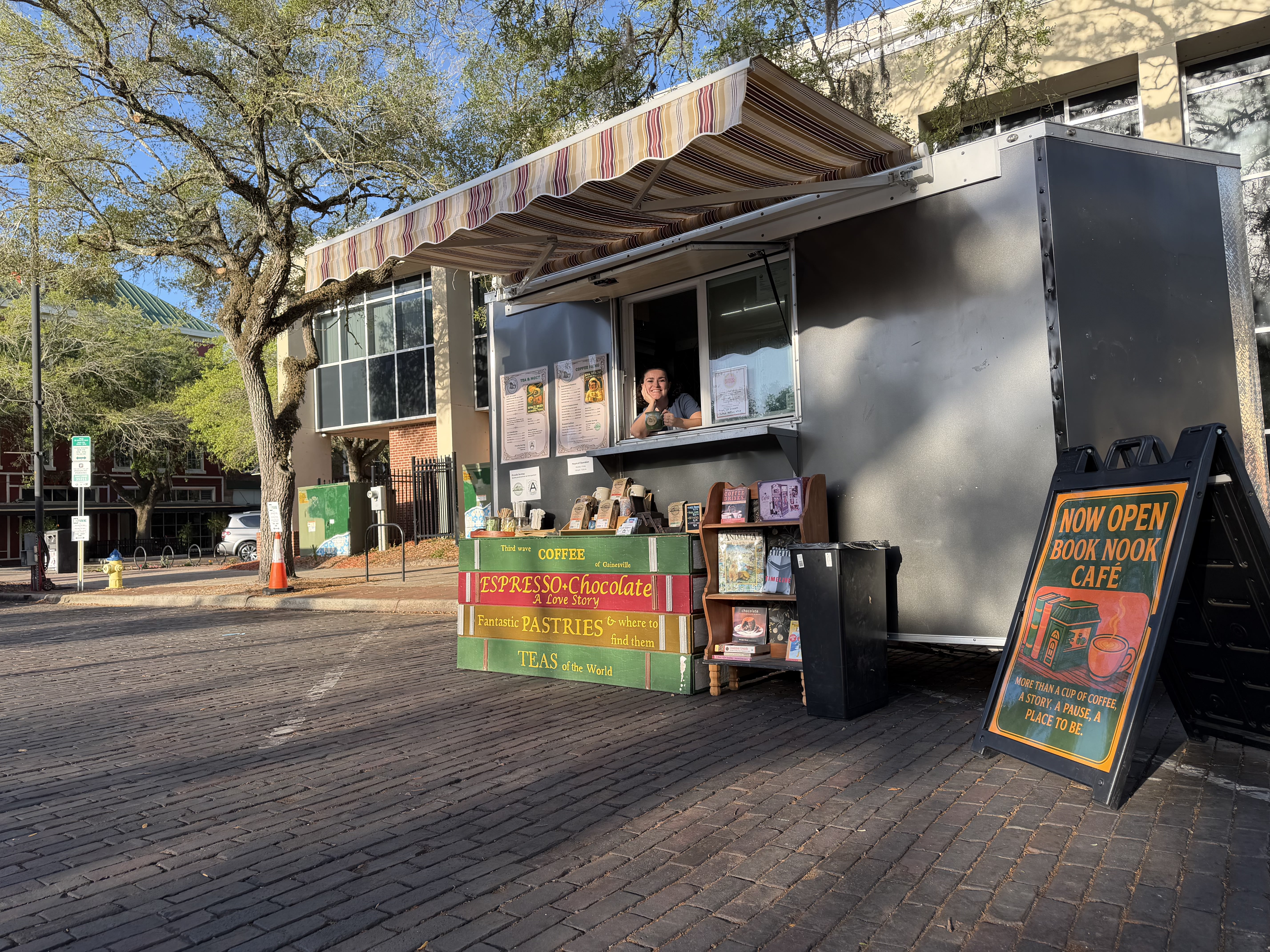 Book Nook Cafe’s coffee trailer with its striped awning, book display, and a smiling team member at the serving window.