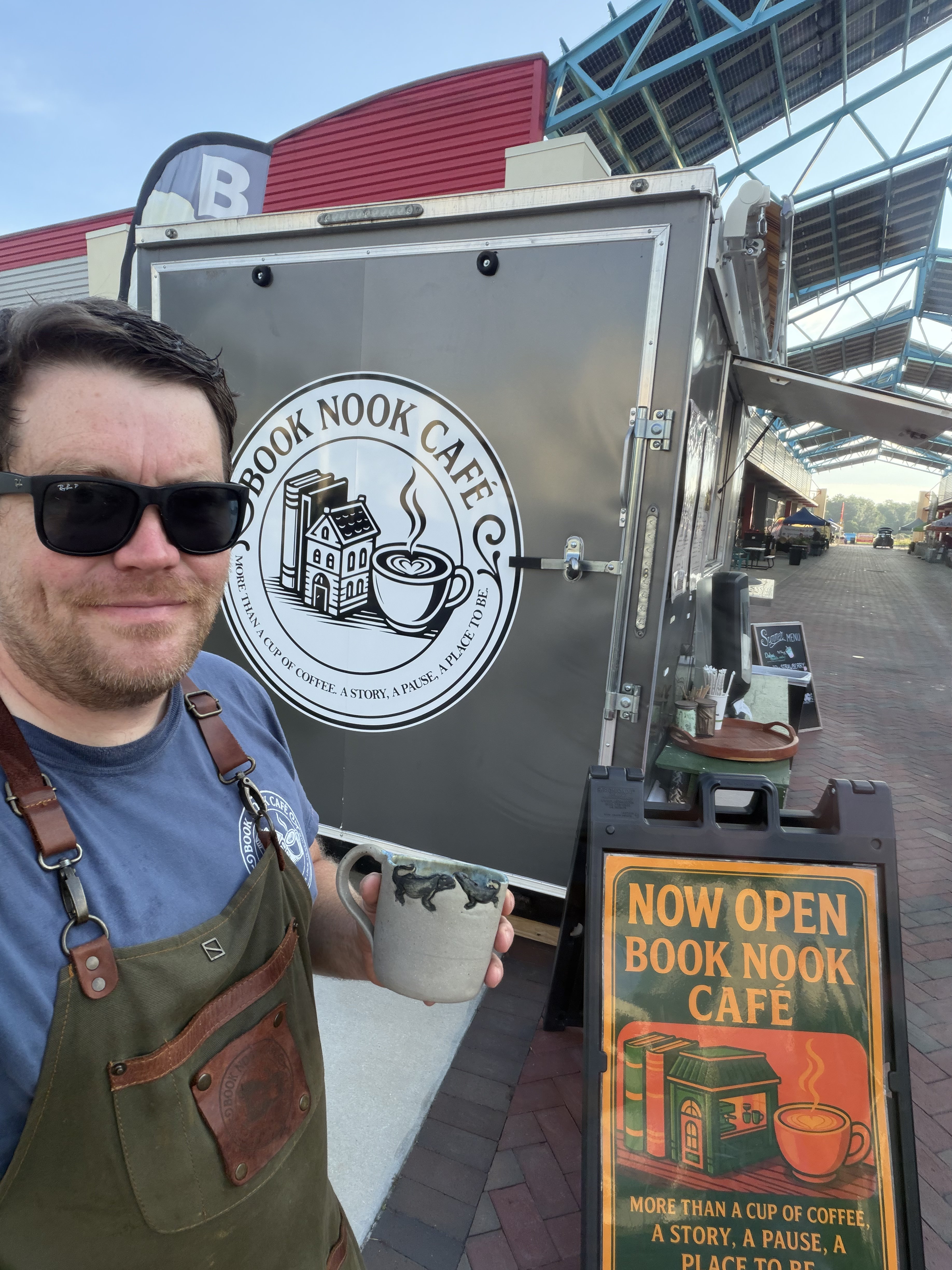 A Book Nook Cafe owner holding a coffee mug beside the trailer and its open sign.